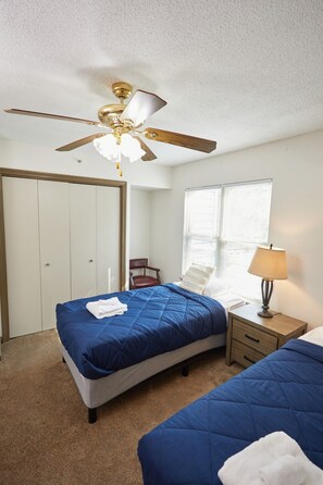 A bedroom with two beds, a ceiling fan, a window with blinds, a chair, and a bedside table with a lamp.