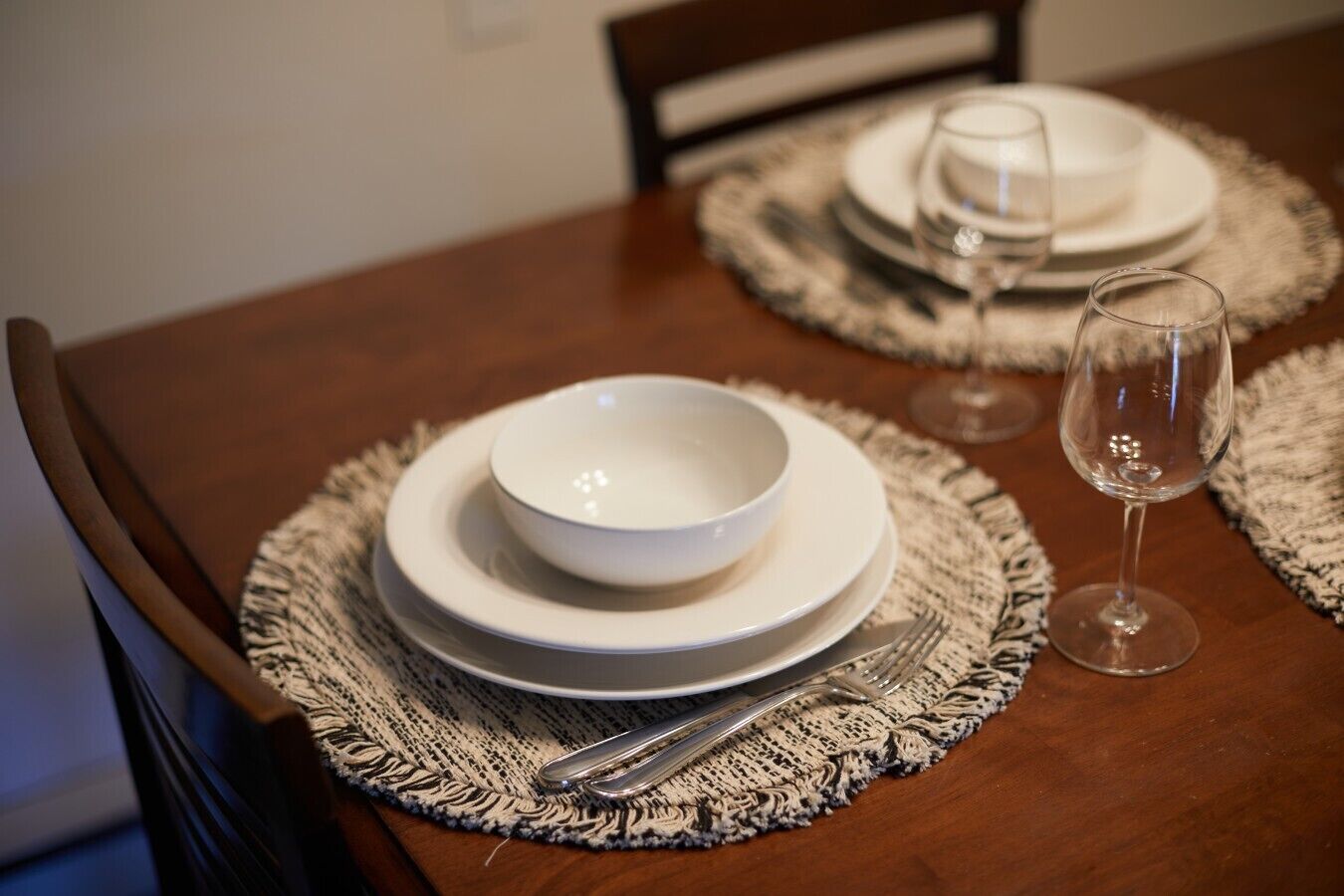 A dining table set with white plates, bowls, and cutlery, and a wine glass on a woven placemat.