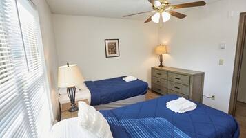 A room with two beds, a ceiling fan, a bedside table with a lamp, a dresser, and a framed picture on the wall.
