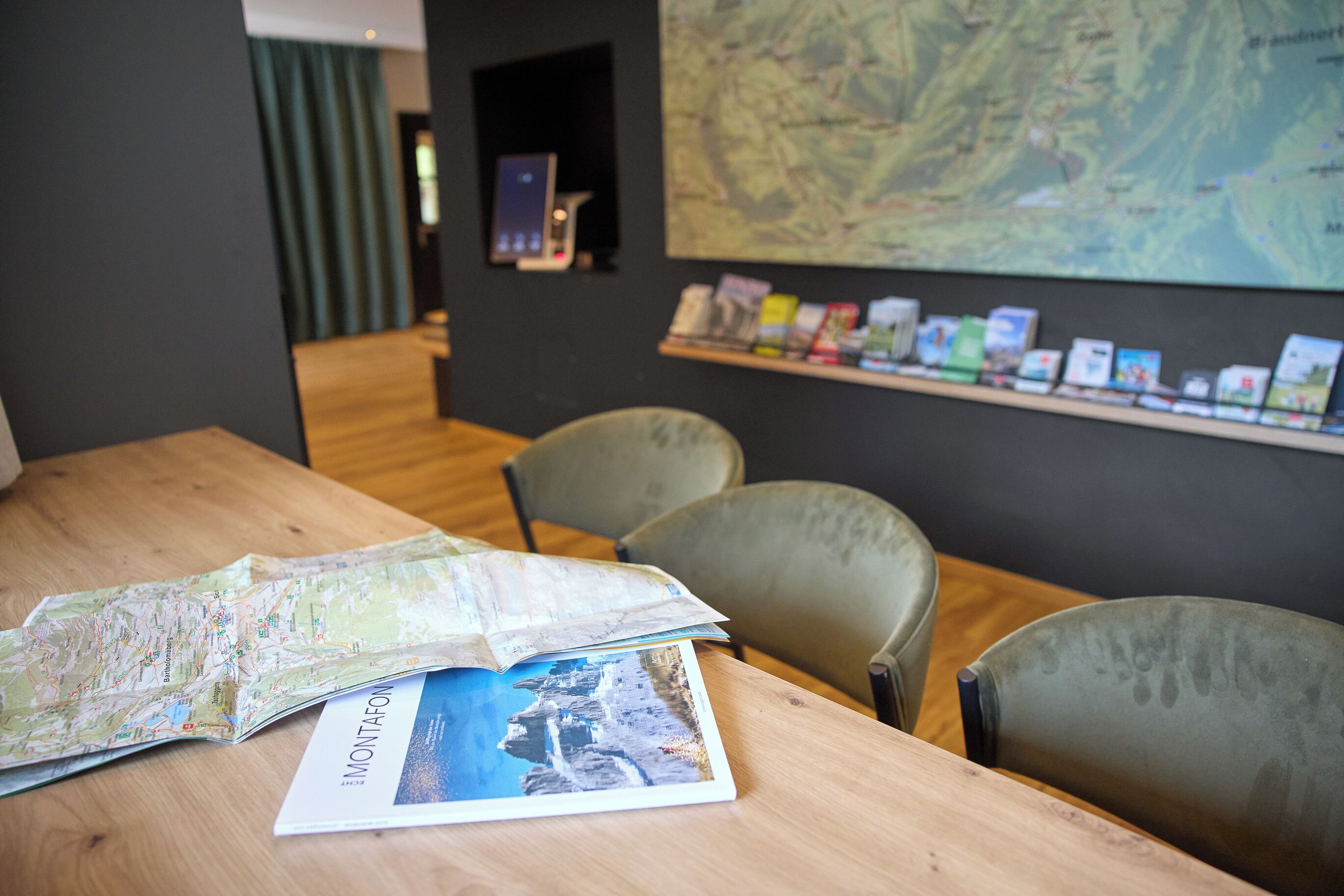 A wooden table with a map and a brochure, a chair, a shelf with books, and a map on the wall.