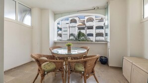 A corner room with a round glass table, wicker chairs, a potted plant, and a view of a building with balconies. - LXR Marysol 2-Bedroom. Apt (Benalmádena)
