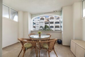 Ein Eckzimmer mit rundem Glastisch, Korbstühlen, einer Topfpflanze und Blick auf ein Gebäude mit Balkonen.