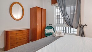 A bedroom with a bed, a wooden wardrobe, a green chair, a window with curtains, and a round mirror on the wall. - LXR Marysol 2-Bedroom. Apt (Benalmádena)