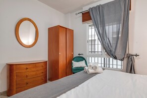 Ein Schlafzimmer mit einem Bett, einem Holzschrank, einem grünen Sessel, einem Fenster mit Vorhängen und einem runden Spiegel an der Wand.