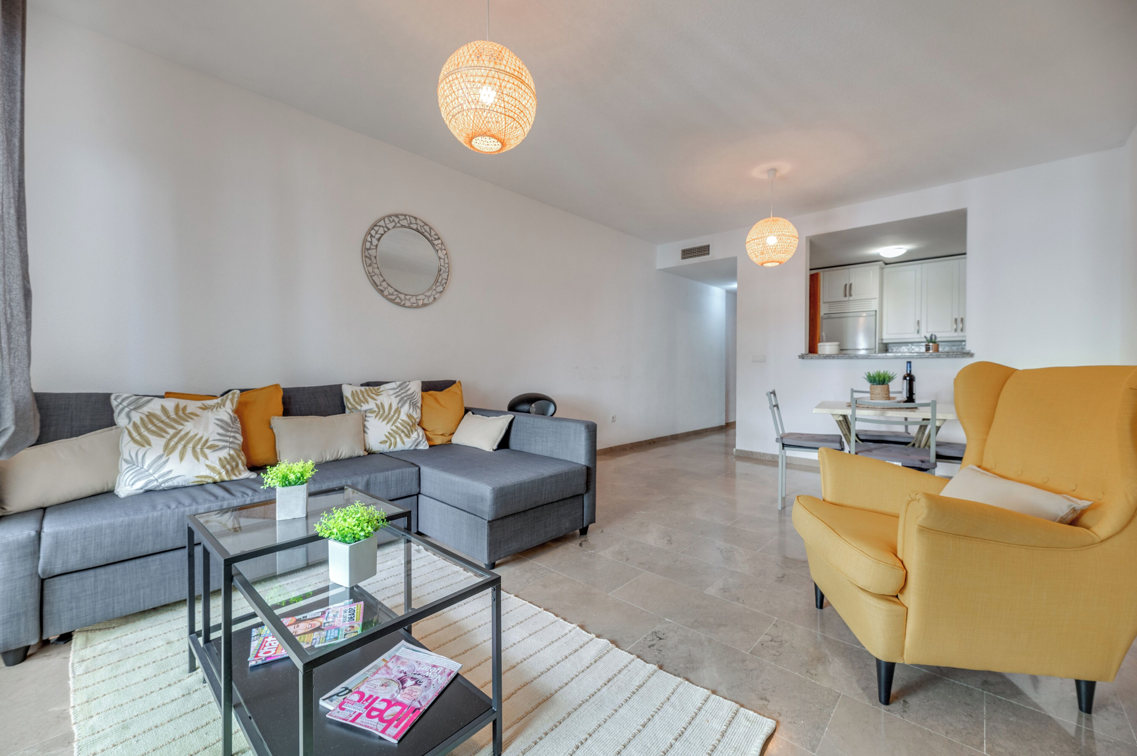 Ein modernes Wohnzimmer mit einem grauen Sofa, einem gelben Sessel, einem Glastisch und einem Essbereich im Hintergrund.