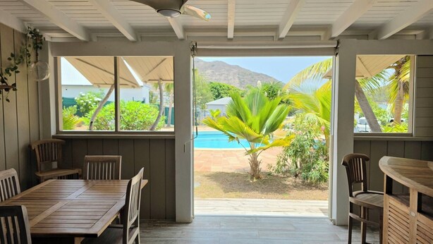 Un patio techado con ventilador de techo, muebles de madera y ventanas amplias que dan a una piscina y plantas tropicales.