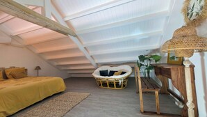 Una habitación con techos altos, una cama, una silla de mimbre, una mesa de madera y una lámpara.