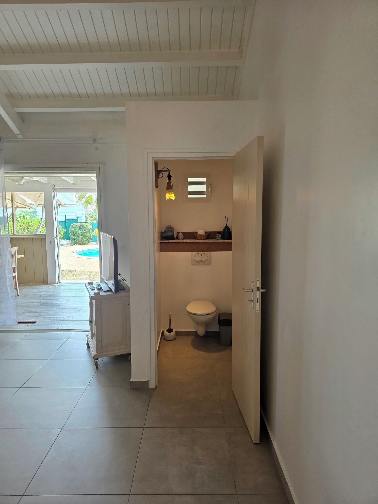 Une salle de bain avec des toilettes, une petite étagère contenant des articles de toilette, un luminaire et une vue sur une piscine à l’extérieur.