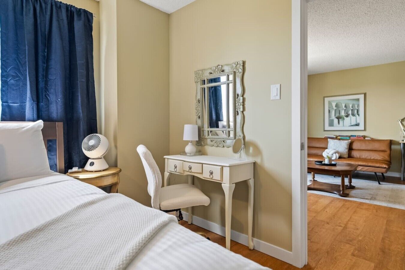 Une chambre d’hôtel avec un lit, un bureau blanc doté d’un miroir, une chaise, une table de chevet avec un ventilateur et un canapé à l’arrière-plan.