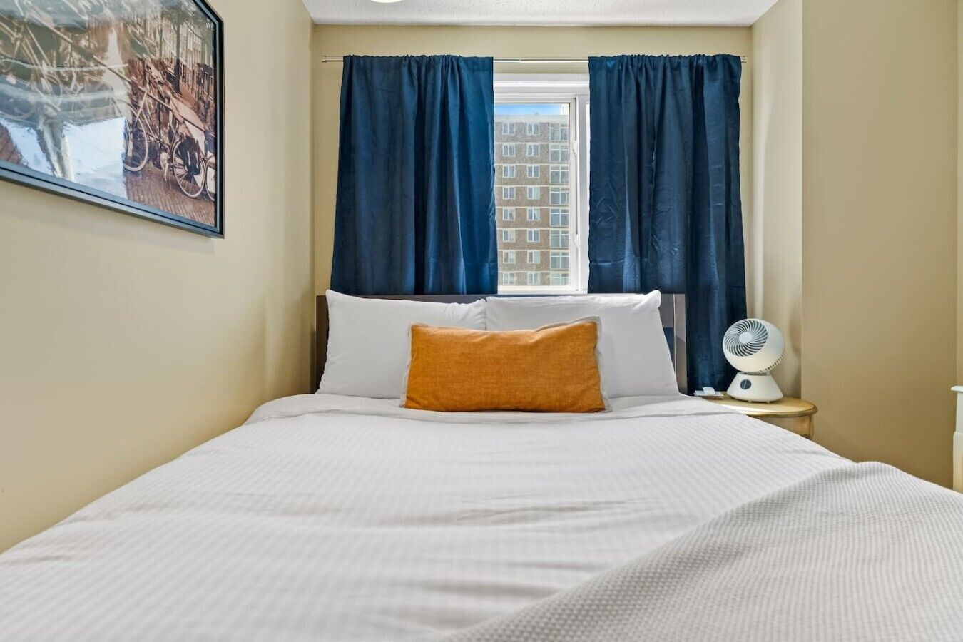 Une chambre d’hôtel avec un lit, deux oreillers, une table de chevet avec un ventilateur et une fenêtre avec des rideaux.