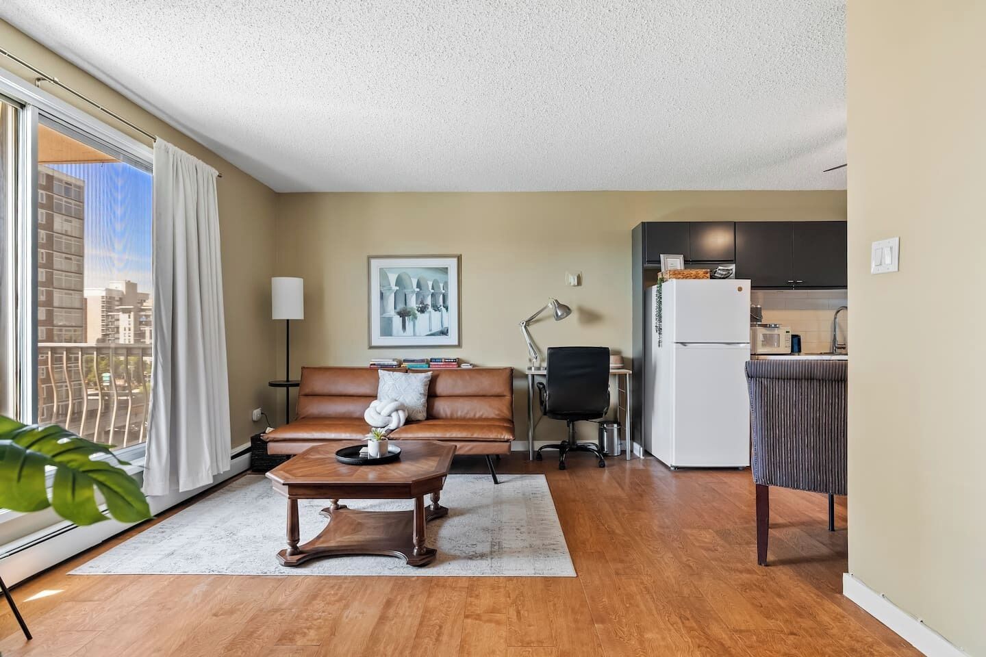 A living room with a sofa, coffee table, and a desk with a lamp. There is a kitchen area with a refrigerator and a microwave.