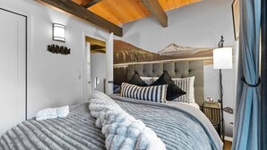 Ein Schlafzimmer mit einem Bett, einem Kopfteil, einem Nachttisch, einer Lampe und einer Wand mit einem Berggebirge.