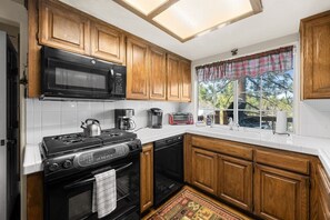 A kitchen with wooden cabinets, a black stove, a microwave, a coffee maker, and a window with a view of trees. - Steps 2 Summit - Walk to Slopes, Pool Table (Big Bear Lake)