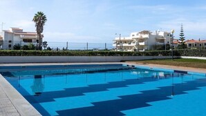 Una piscina con un diseño geométrico de azulejos, rodeada por un seto bajo y una hilera de edificios blancos con balcones.