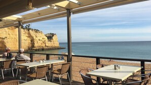 Un área de comedor al aire libre con mesas y sillas, con vistas a una playa y acantilados.