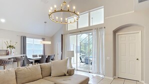 Un salon moderne avec un canapé d’angle, un lustre, un coin repas et une porte-fenêtre coulissante donnant sur une terrasse extérieure.