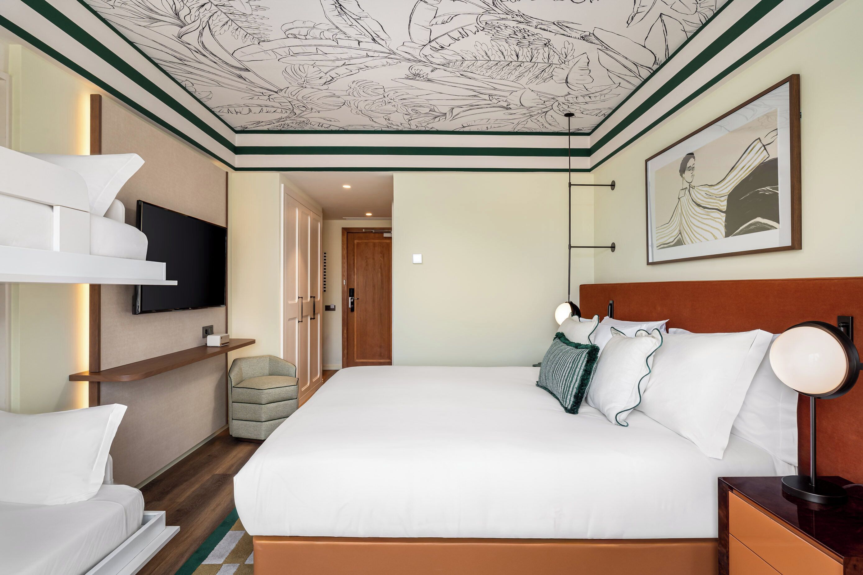 A hotel room with a large bed, a television mounted on the wall, a framed artwork above the bed, and a wooden headboard.