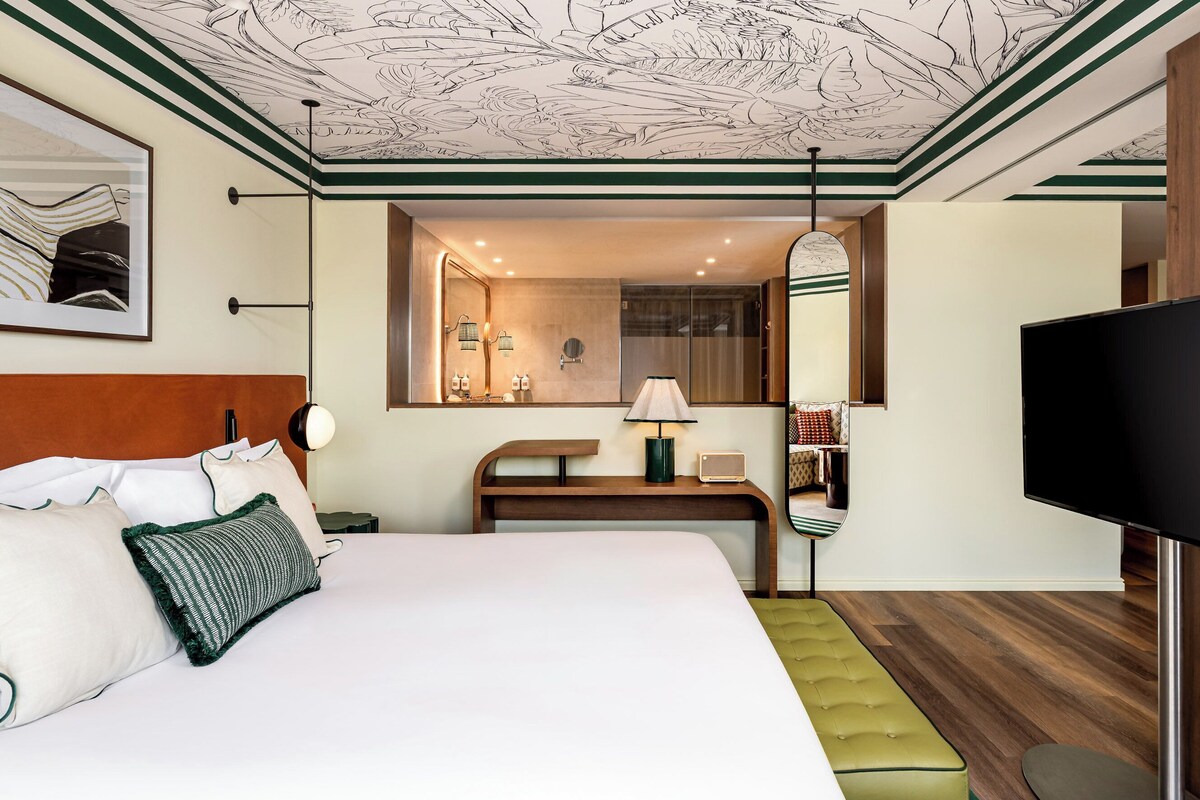 Habitación de hotel con cama, mesita de noche, lámpara, espejo y televisión.