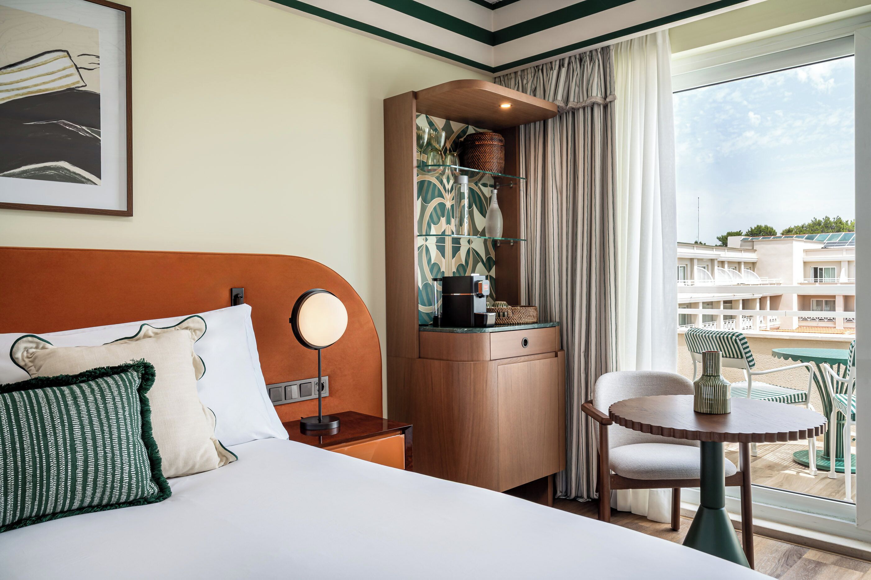 A hotel room with a bed, a bedside table with a lamp, a small table with a vase, a chair, and a view of the outside.