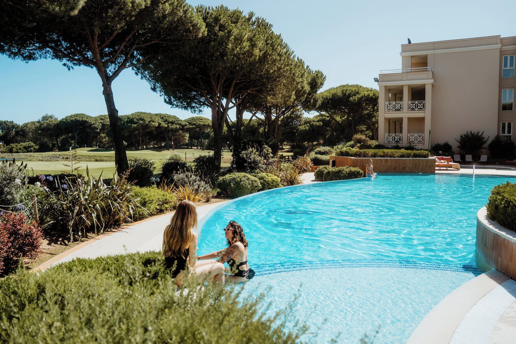 A swimming pool with a clear blue water, surrounded by greenery and a building with a balcony.