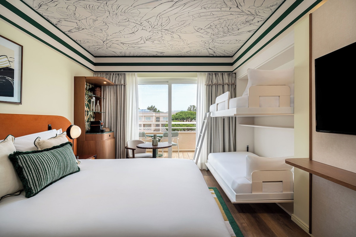 Habitación de hotel con cama, una mesita con sillas, televisión y balcón con vistas.