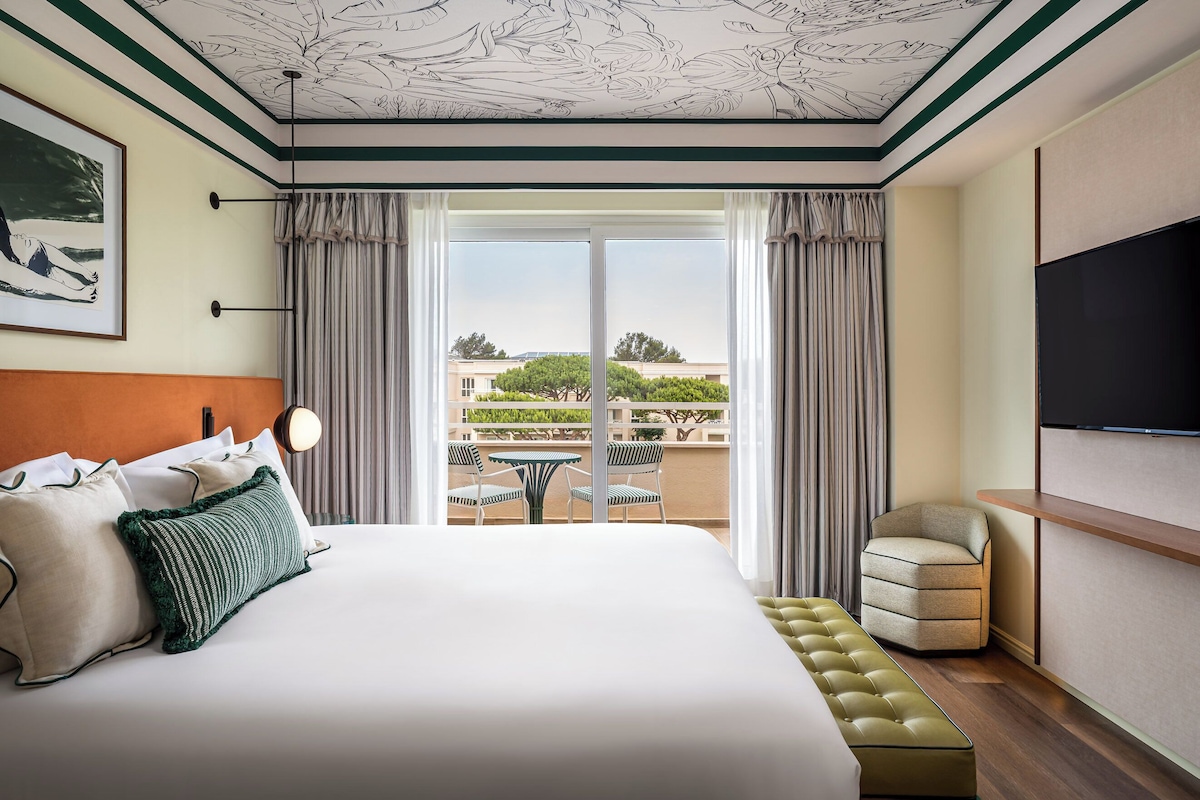 Habitación de hotel con una cama grande, televisor, balcón con mesa y sillas, y vistas del entorno.