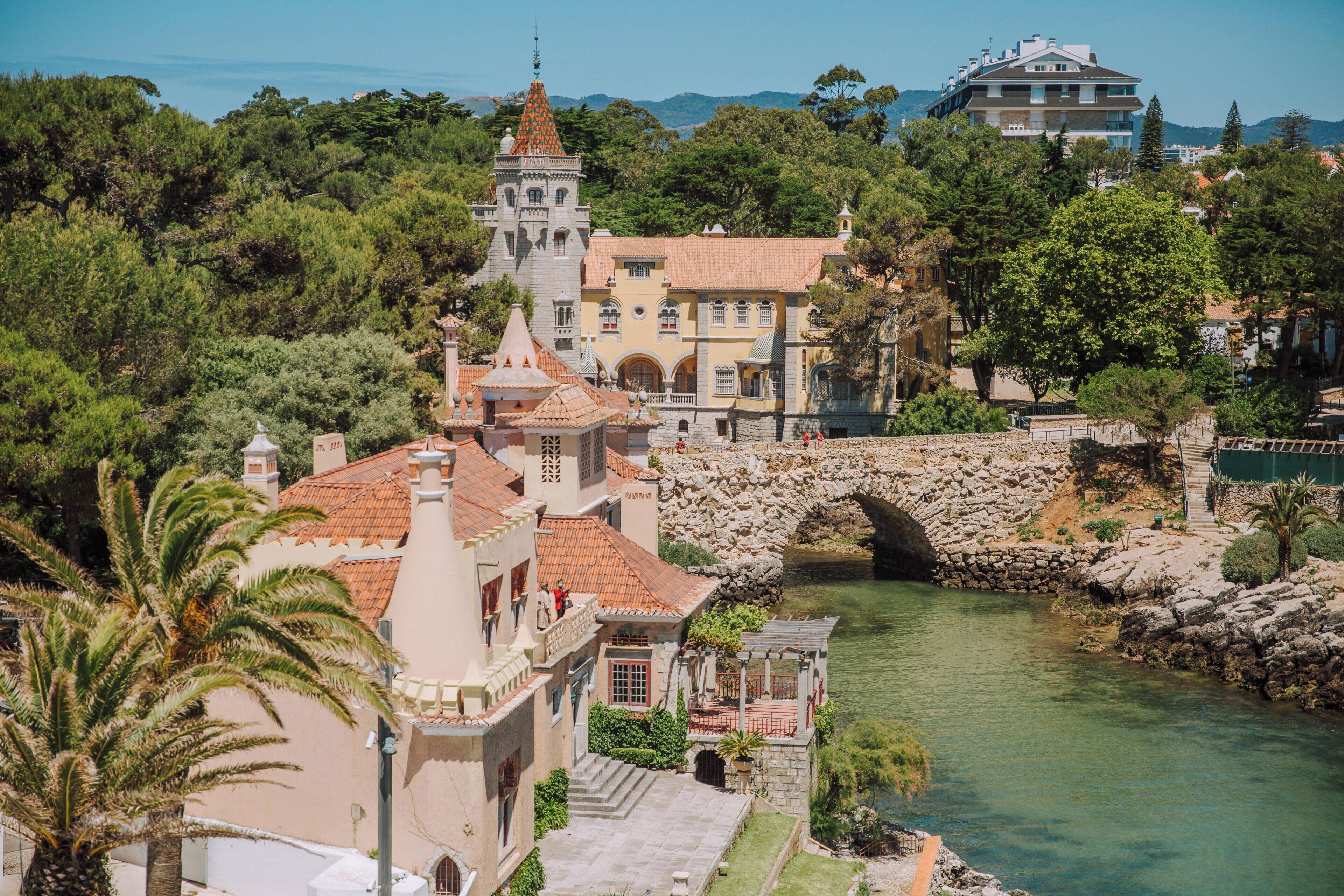 A historic building with a stone bridge over a river, surrounded by lush greenery and a clear sky.