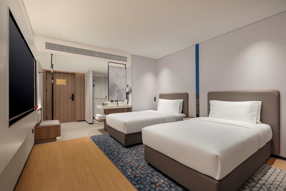 A hotel room with two beds, a TV, a bathroom, and a patterned rug.