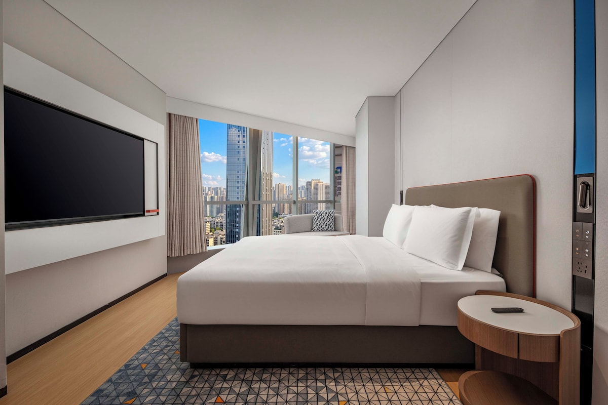 A modern hotel room with a large bed, a flat-screen TV, a round side table, and a city view.