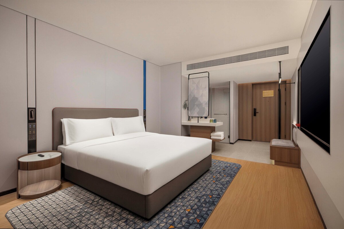A modern hotel room with a large bed, a bedside table, a small bedside table, a mirror, a wardrobe, and a TV.