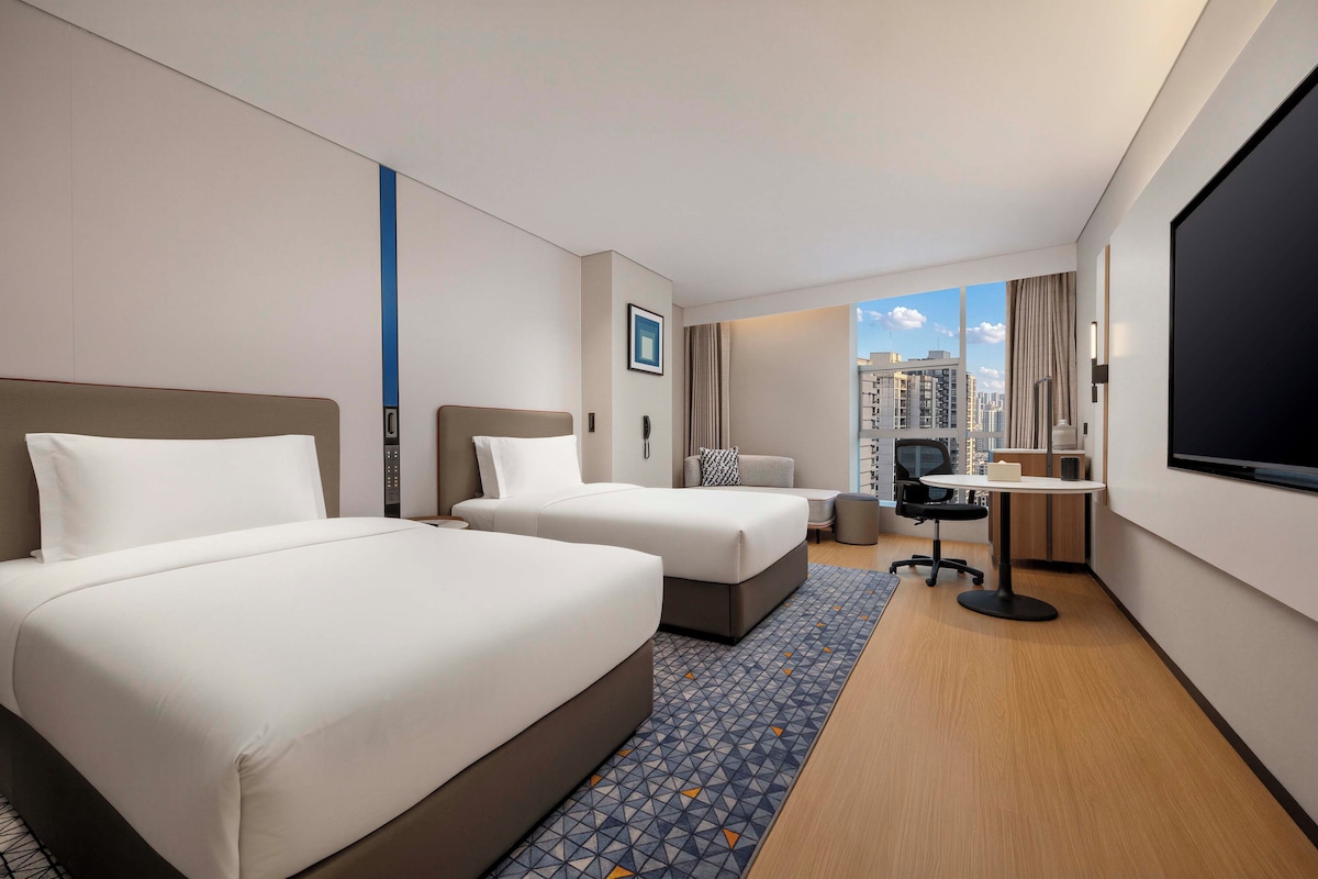 A hotel room with two beds, a desk, a chair, and a large TV screen.
