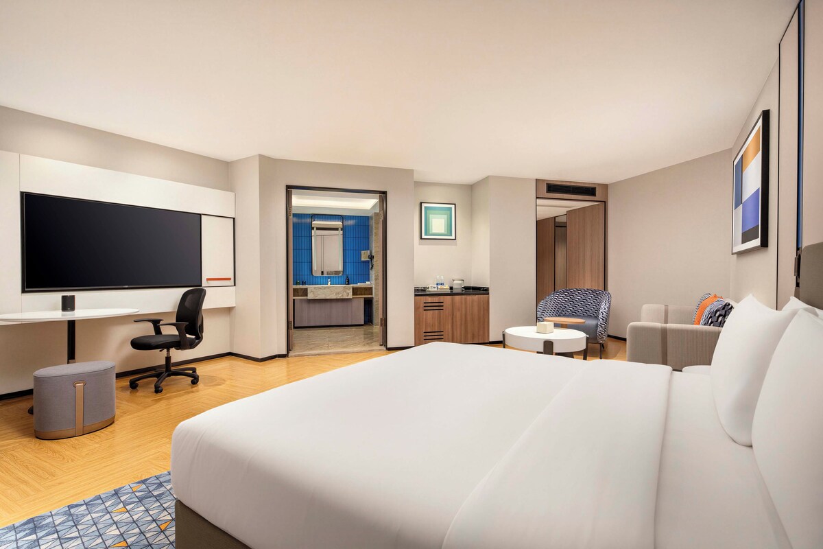 A hotel room with a large bed, a desk with a computer, a chair, a small table, and a TV.
