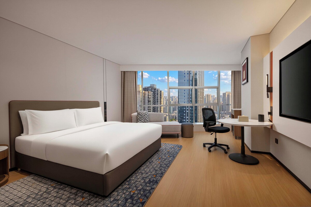 A hotel room with a large bed, a desk, a chair, a TV, and a view of the cityscape.