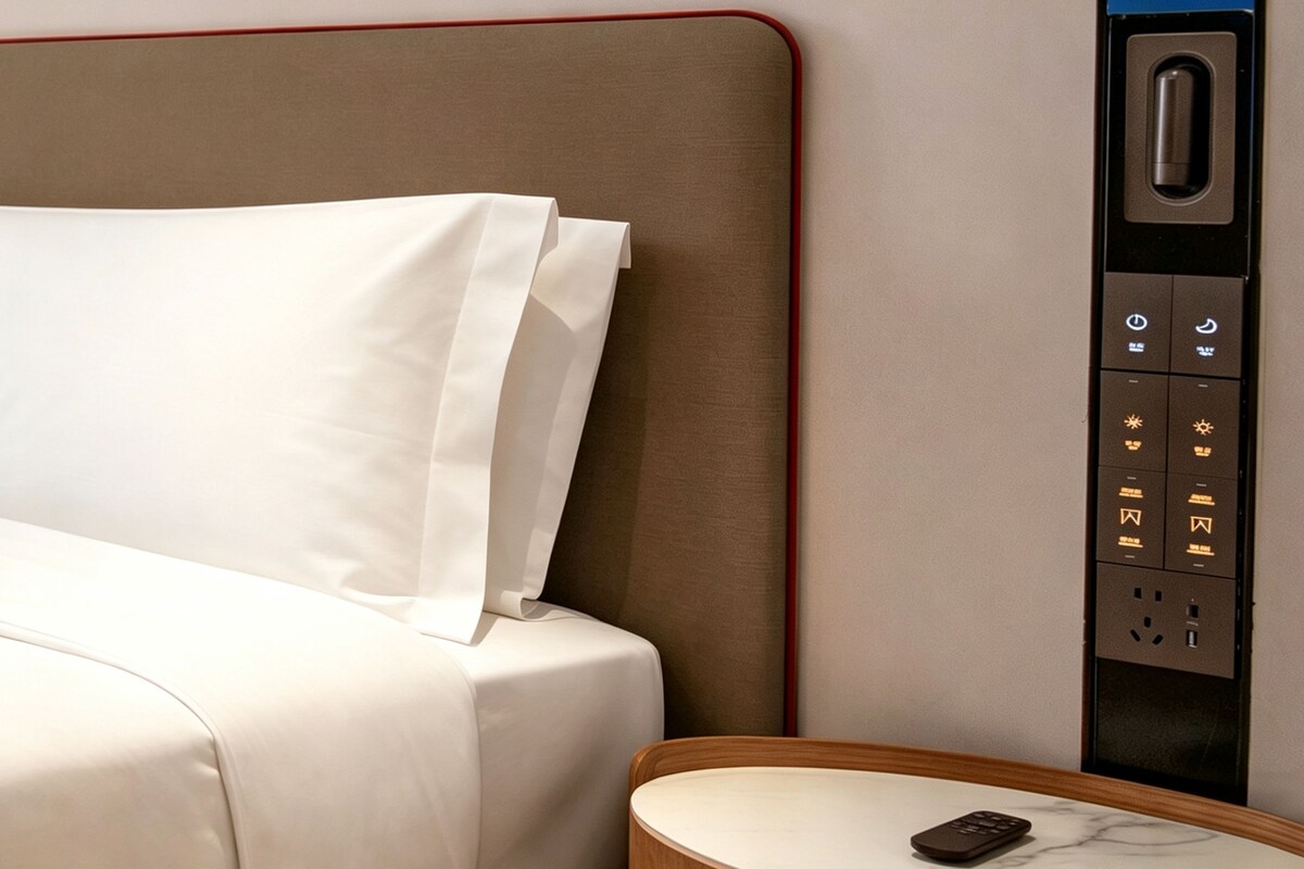 A hotel room with a bed, a nightstand, and a wall-mounted control panel with various buttons.
