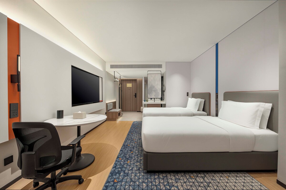 A modern hotel room with a flat-screen TV, a desk, a chair, two beds, and a bathroom with a shower.