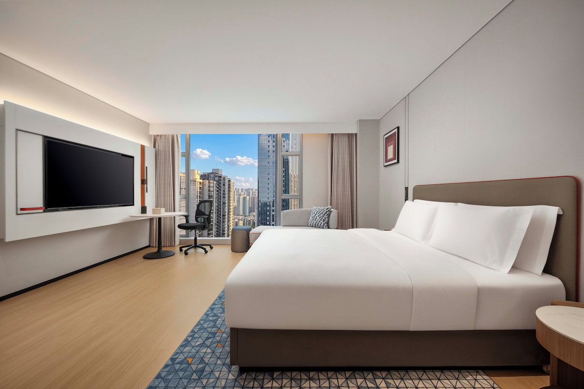 A modern hotel room with a large bed, a flat-screen TV, a desk, and a view of the cityscape.