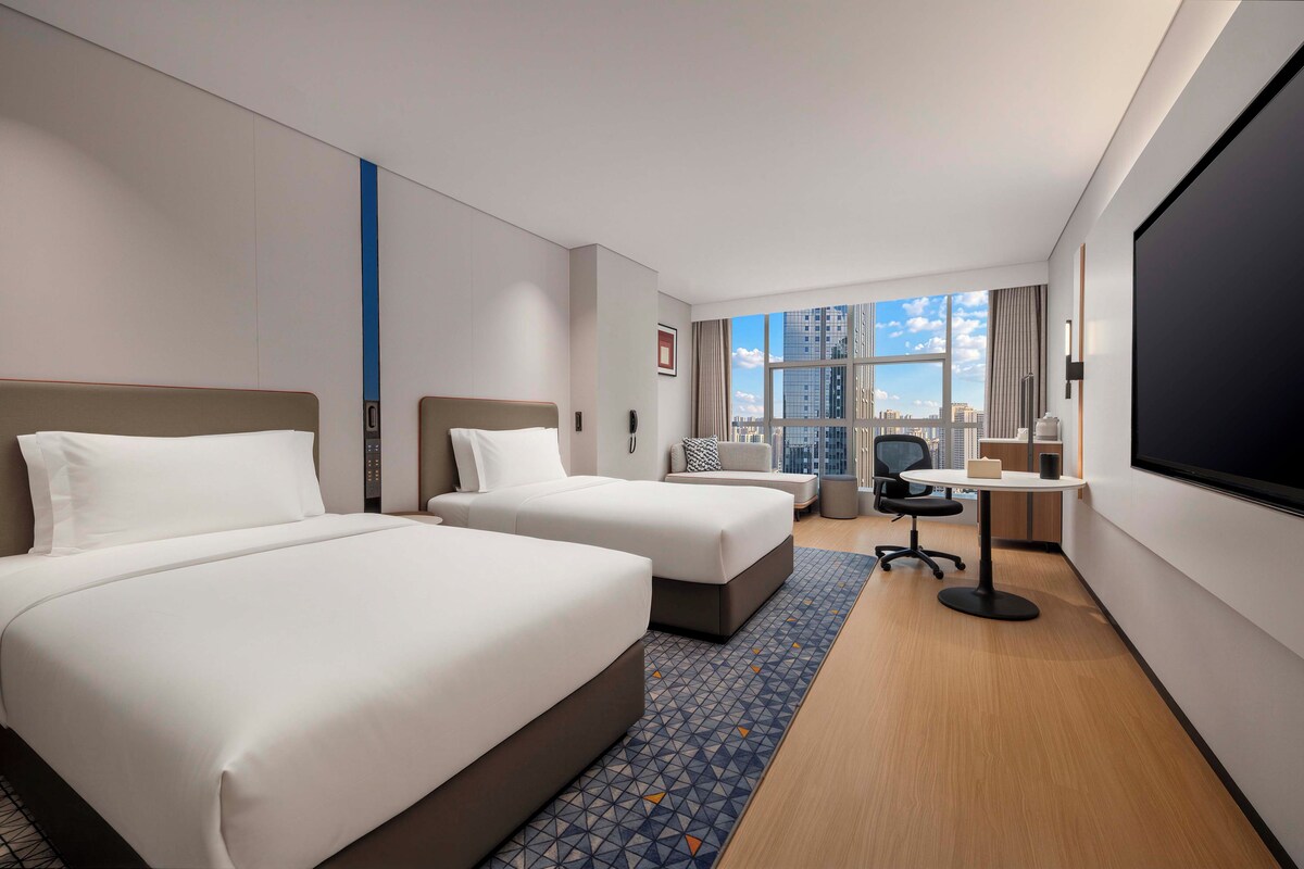 A hotel room with two beds, a desk, a chair, a TV, and a large window with a city view.