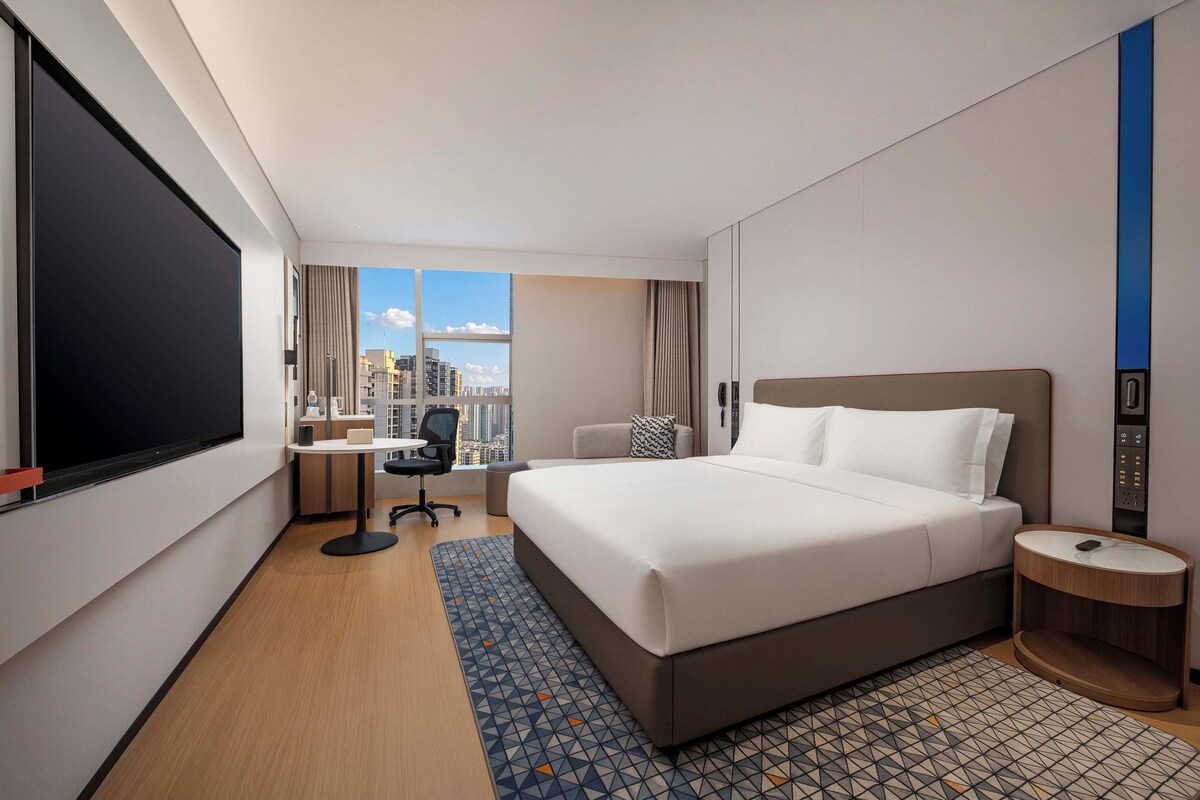 A hotel room with a large bed, a desk, a chair, a small table, a phone, and a window with a city view.