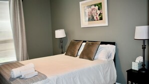 Een keurig opgemaakt bed met kussens, een bedlampje, een ingelijste foto aan de muur, en een raam met gordijnen.