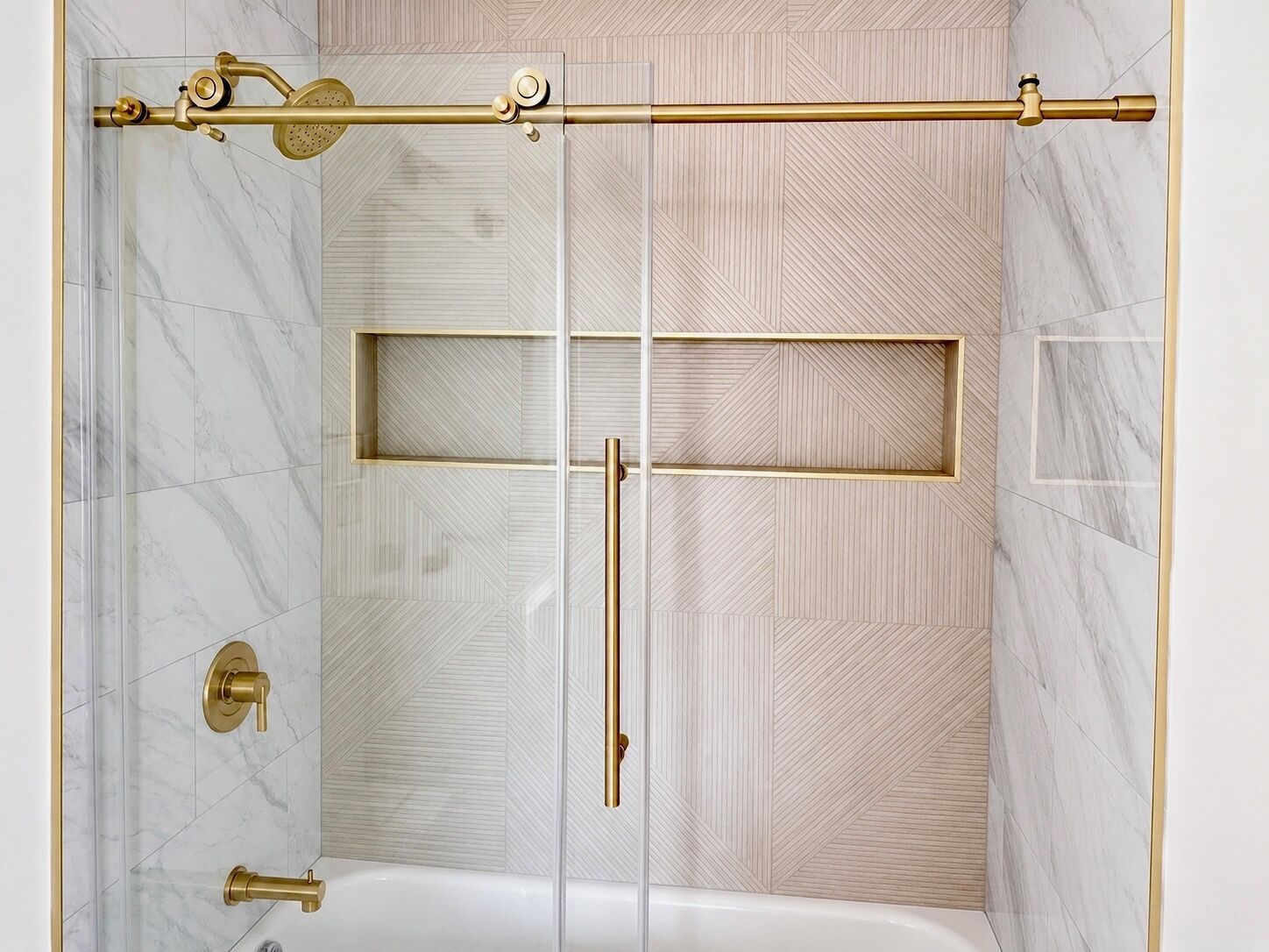 A shower with a gold showerhead and control panel, a glass door, and a marble-patterned wall.