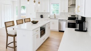 A modern kitchen with white cabinetry, a central island, a dining area with chairs, and a large window providing natural light. - Brand New 2026 Villa, 5 min Walk to the Beach, King Canopy Suite, Sleeps 16 (Keansburg)