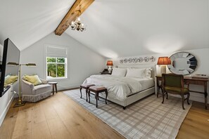 Een slaapkamer met een schuine kap, een bed met witte beddengoed, een nachtkastje met een lamp, een stoel, een ronde spiegel en een raam met uitzicht op groen.