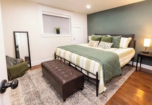 A bedroom with a bed, a bench, a bedside table, a lamp, a window, and a mirror.