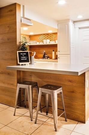 A kitchen with a wooden accent wall, a white countertop, two stools, and a sign reading 'Welcome to Tramway Basecamp.' - Tramway Base camp: Your home away from home close to the best recreation in Utah (Sandy)