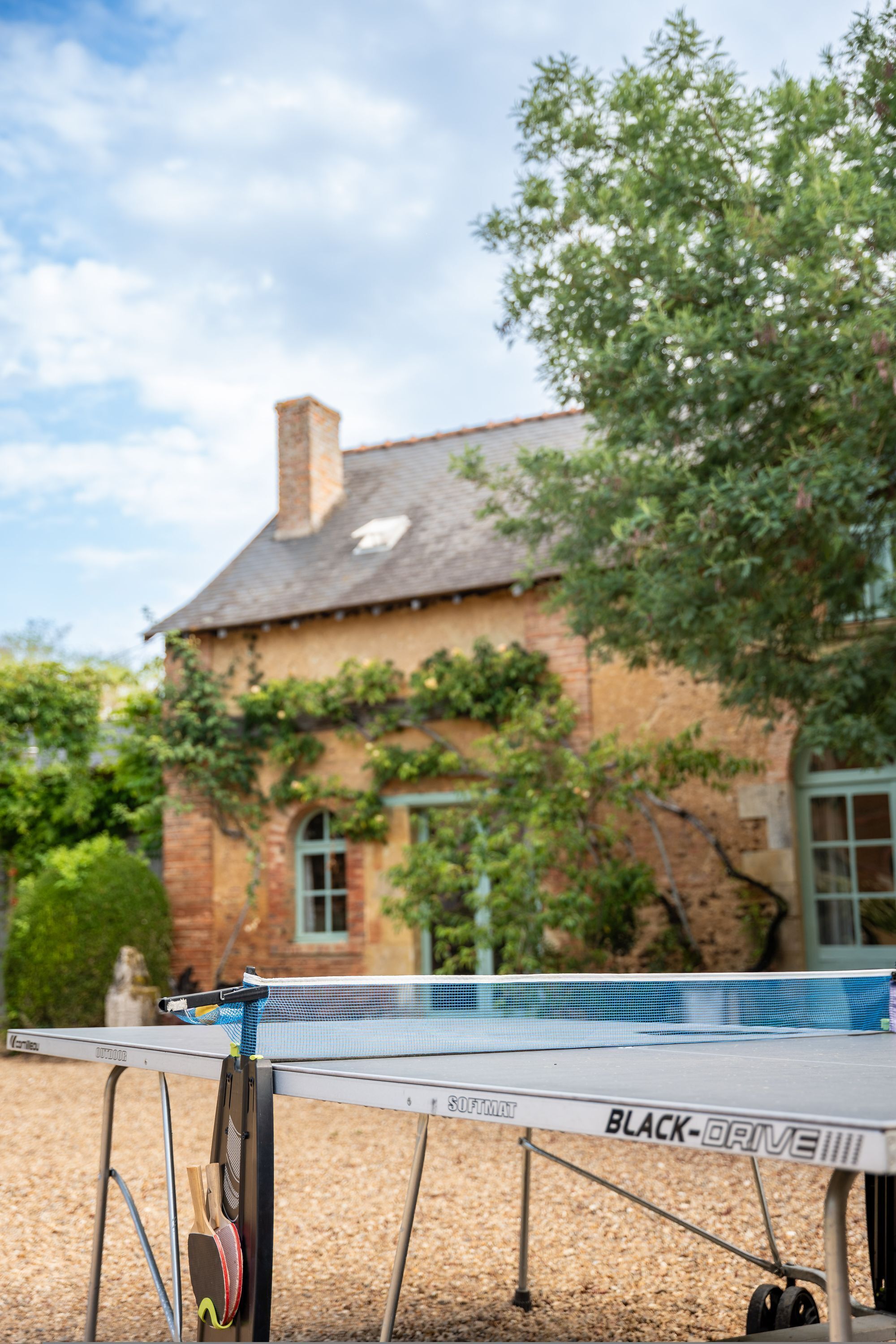 A table tennis table with a net, positioned outdoors with a brick building and greenery in the background.