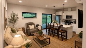 A modern living room with a sofa, armchair, and coffee table, a dining area with chairs, a kitchen with white cabinets and appliances, and a large window with a view of greenery.