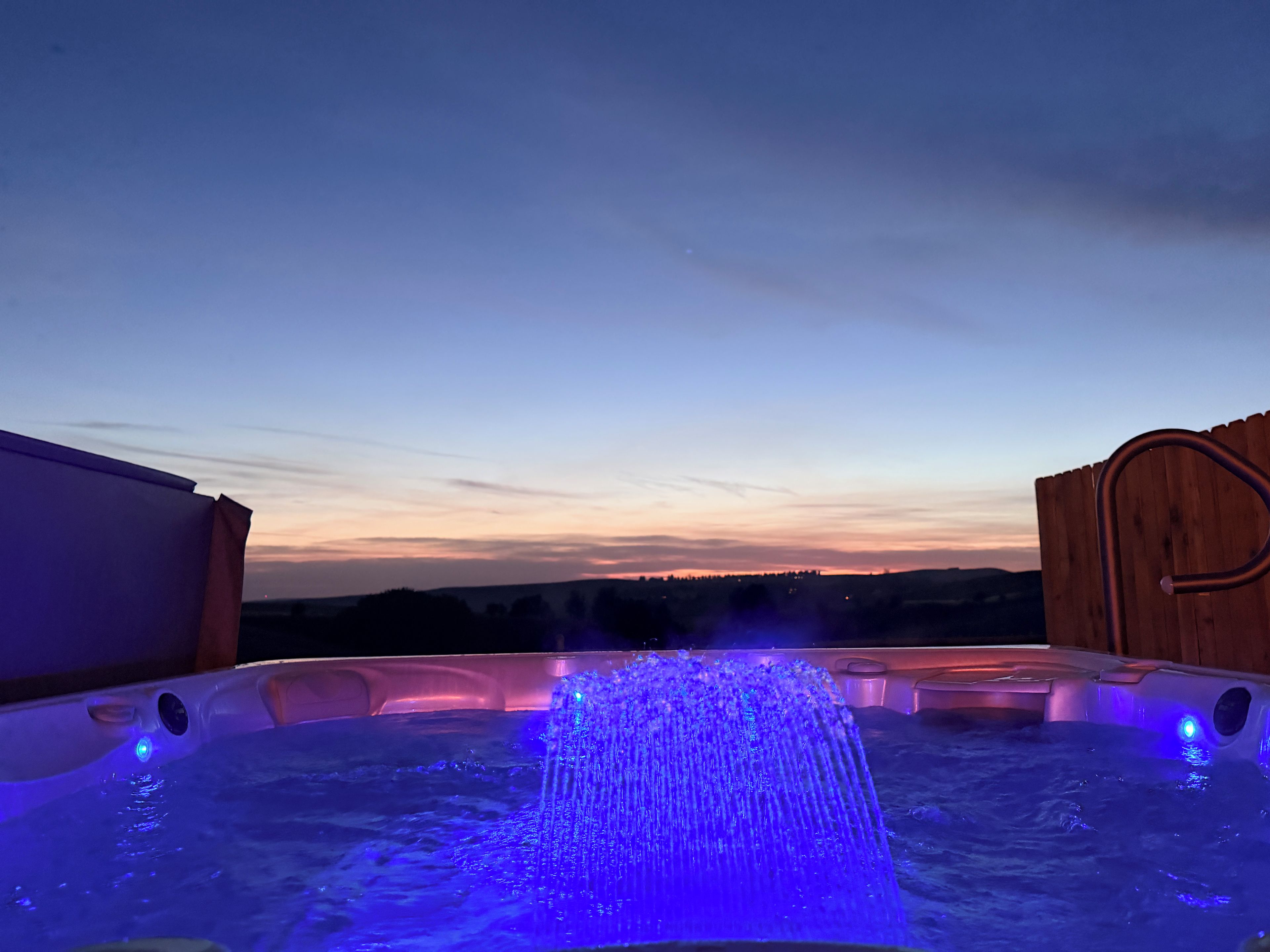 A hot tub with a water fountain feature, surrounded by a wooden deck, with a view of the landscape in the background.
