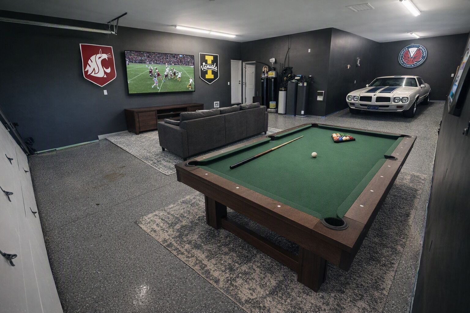 A pool table with a green felt surface, a sofa, a television displaying a sports game, and a classic car in the background.