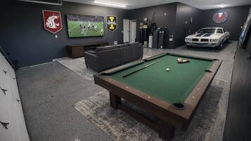 A pool table with a green felt surface, a sofa, a television displaying a sports game, and a classic car in the background.