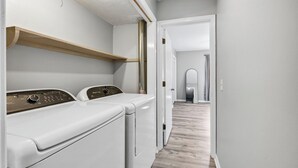 A laundry room with two front-loading washers and dryers, a wooden shelf above the dryers, and a hallway leading to a door. - Southern Oasis-Modern Comfort, Minutes from Downtown (Columbia)